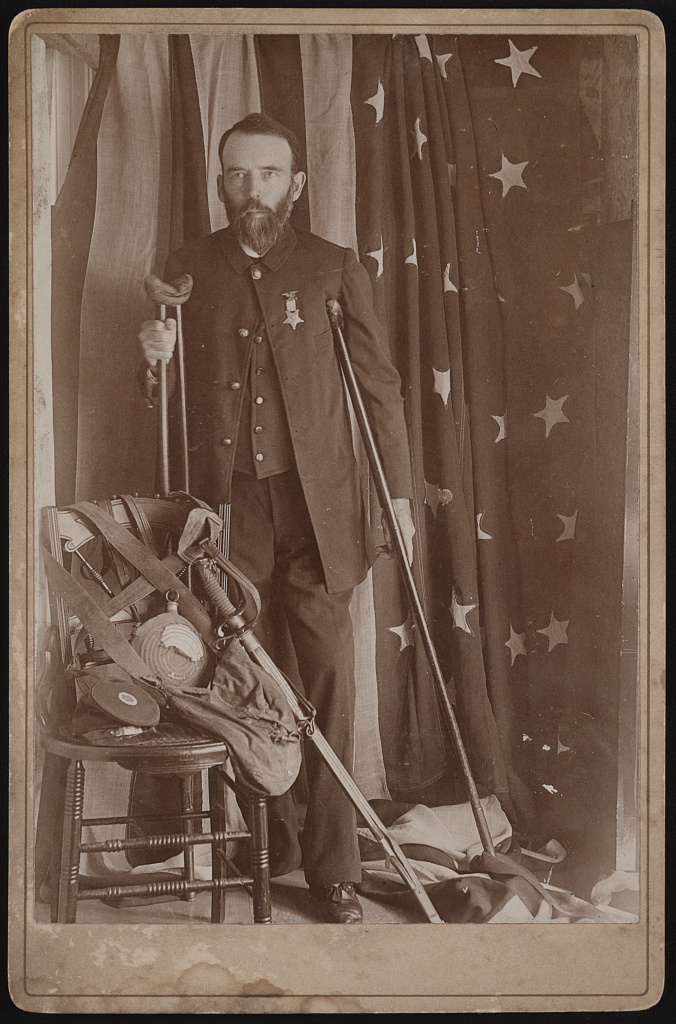 Henry A. Seaverns in uniform with sword, canteen, and other artifacts, standing on crutches in front of American flag