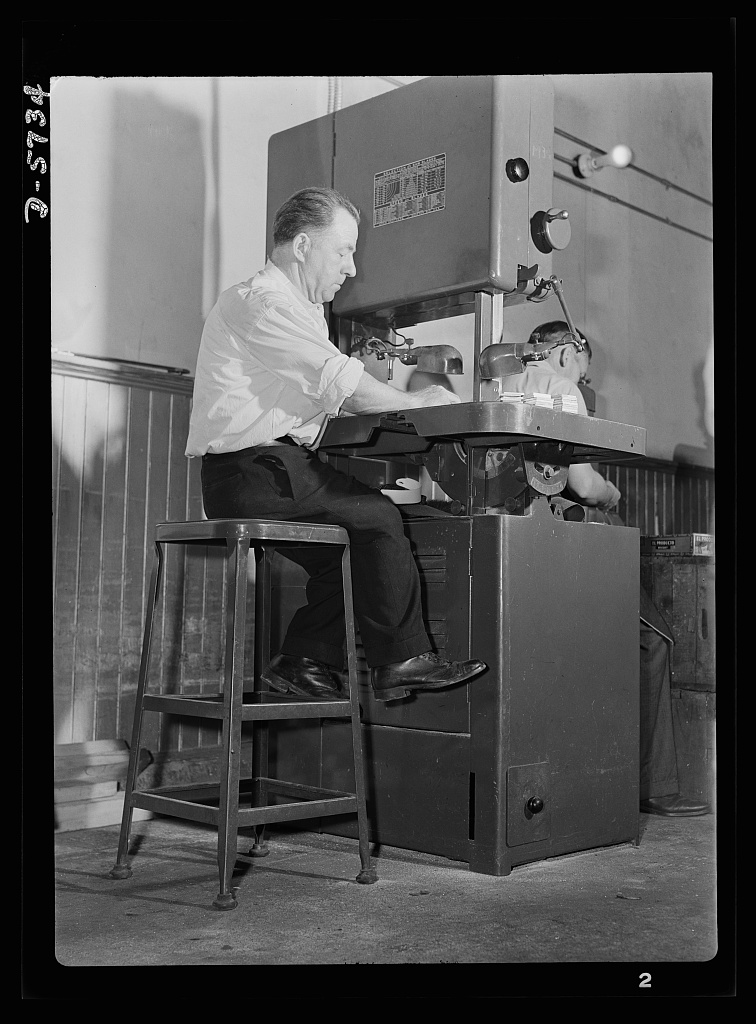 a man operating a massive saw while sitting on a stool, his legs too short to reach the ground