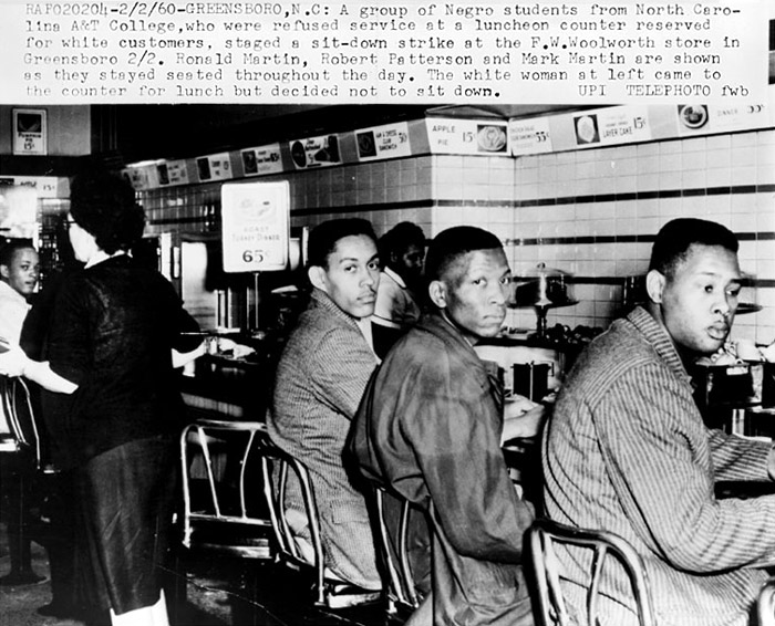 Three young black men on chairs at a service counter look over their right shoulders with serious faces. Manual typewriter text visible above describes the scene, noting that a white woman seen in the background is walking away having decided not to sit and have lunch. 