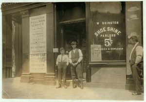 City sidewalk with two boys in shirtsleeves in doorway of storefront, "Shoe Shine Parlor 5 cents for ladies and gentlemen"