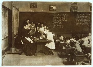 A classroom with students working at desks, teacher in long dress seated at a table next to chalk boards, surrounded by a group of 6 girls of varied heights, using her hands as she explains something.