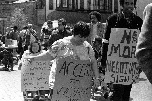 Protesters, black and white, walking and in wheelchairs, carry signs demanding "Access to Work" and "Sign 504"
