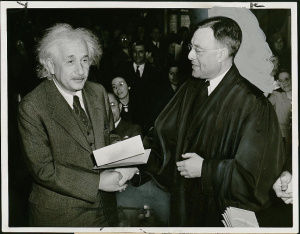Albert Einstein holds papers and shakes hands with another man.