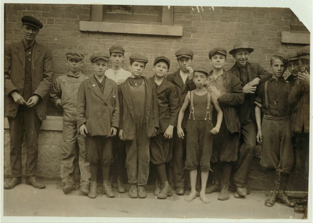 Photo of several boys dressed for factory  work. A younger barefoot boy smokes a pipe, probably a prank.