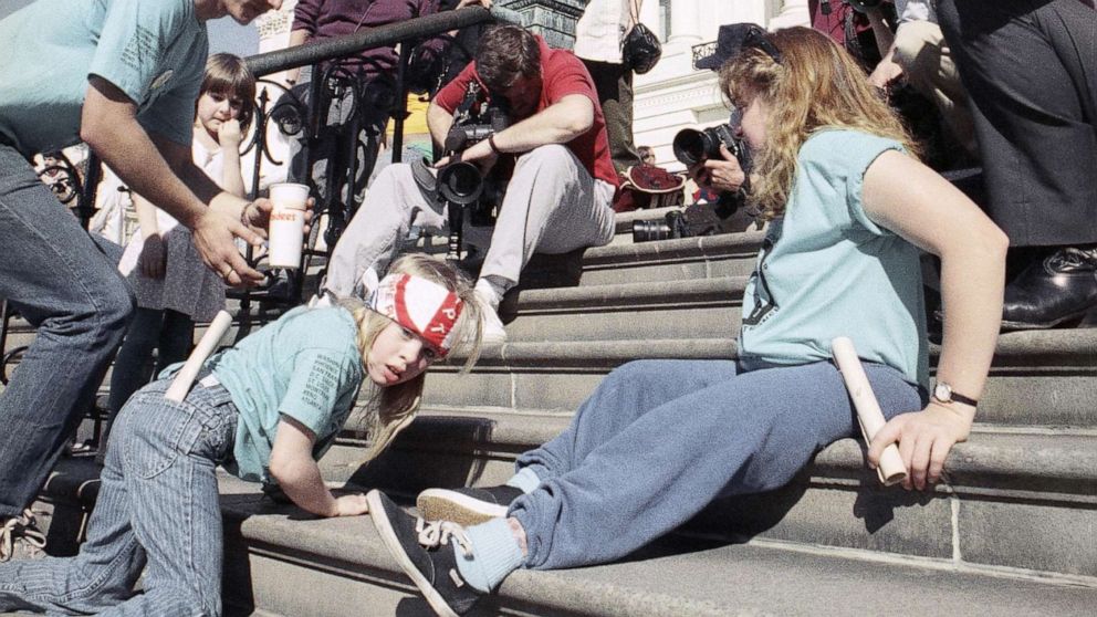 Color Photo shows girl sitting on steps using her arms to lift her torso to the step behind her as she is surrounded by other protesters and members of the press