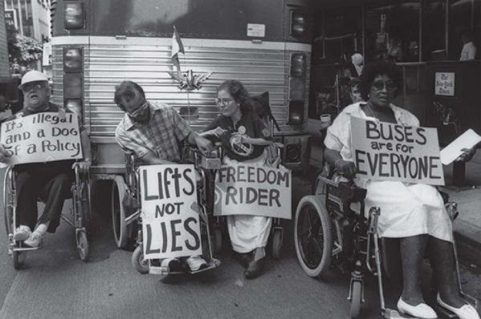 People in wheelchairs blocking path of bus, holding signs, "Buses are for Everyone." 