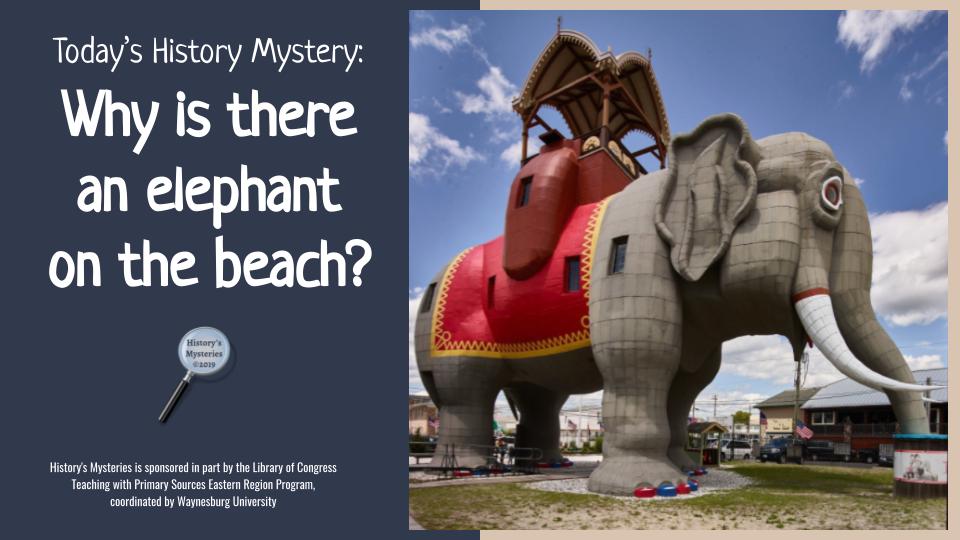 Today's Mystery Why is there an elephant on the beach? - huge grey elephant structure towers over nearby buildings, sandy grass in foreground and blue sky behind