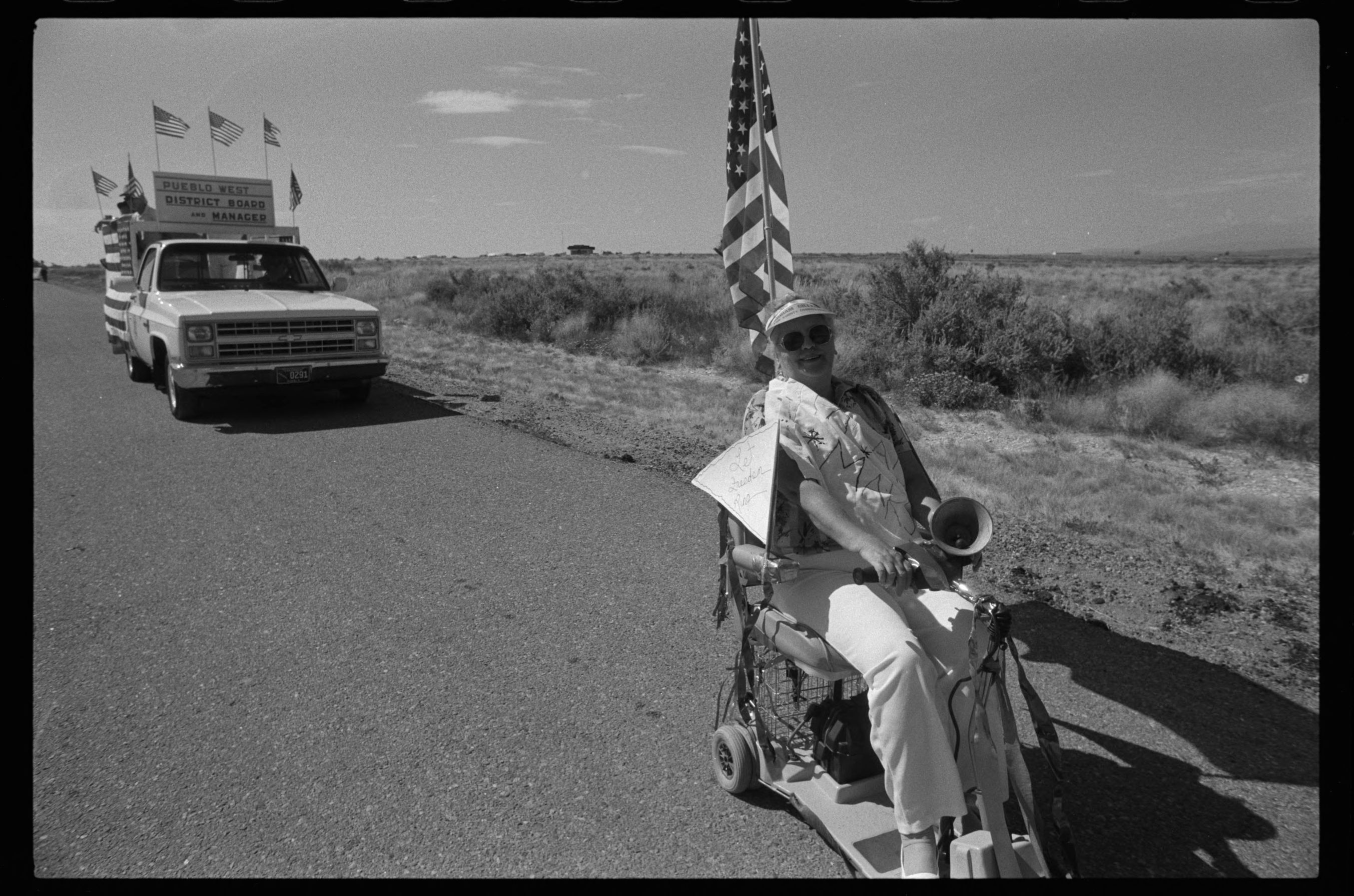 On an open road, a woman steers her motorized scooter, bullhorn on her lap, leading a pickup truck with many US flags driving behind.