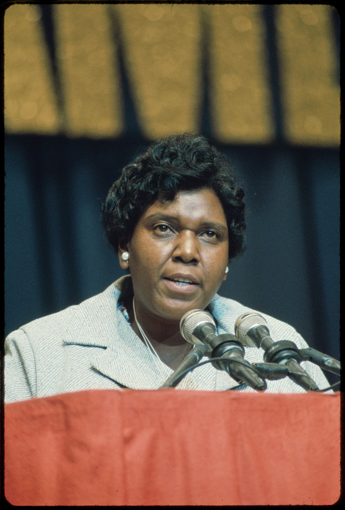 A photograph of Congresswoman Barbara Jordan at a podium speaking into microphones. Congresswoman Jordan is a middle aged African American woman who is wearing a gray coat, large pearl earrings, and has short, wavy, black hair.