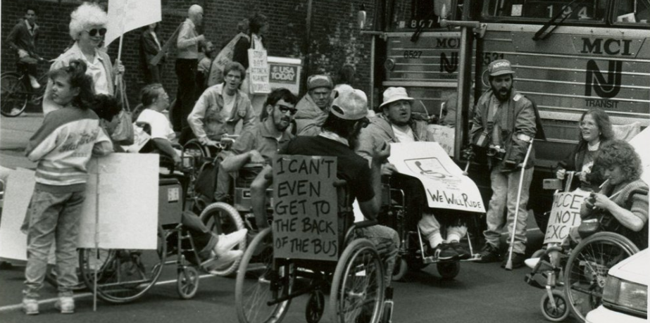 A group of ADAPT Activists protest for accessible public transportation in the street outside a transit center. The activists are mostly wheelchair users. They hold or wear handmade signs, including one saying, "I can't even get to the back of the bus."