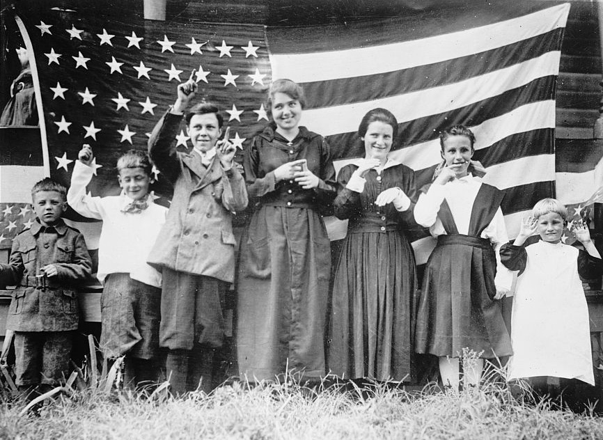A photograph of a group of children in front of a large American flag. The children are dressed in early 20th century clothing and have their hands up in the middle of signing the national anthem.