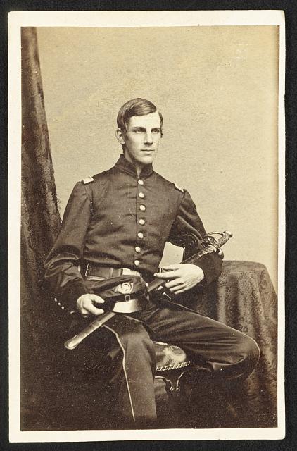 Photo of a clean-shaven young officer in Union uniform, seated, with a cap bearing the brass horn of infantry. He cradles an officers sword in his lap and looks aside from the camera.  
