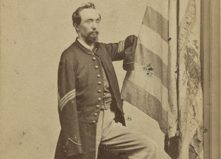 A bearded young man stands in an unbuttoned sergeant's jacket, showing the three chevrons of his rank. Both his sleeves are empty. His right upper arm is raised, resting on the staff of a tattered United States flag. 