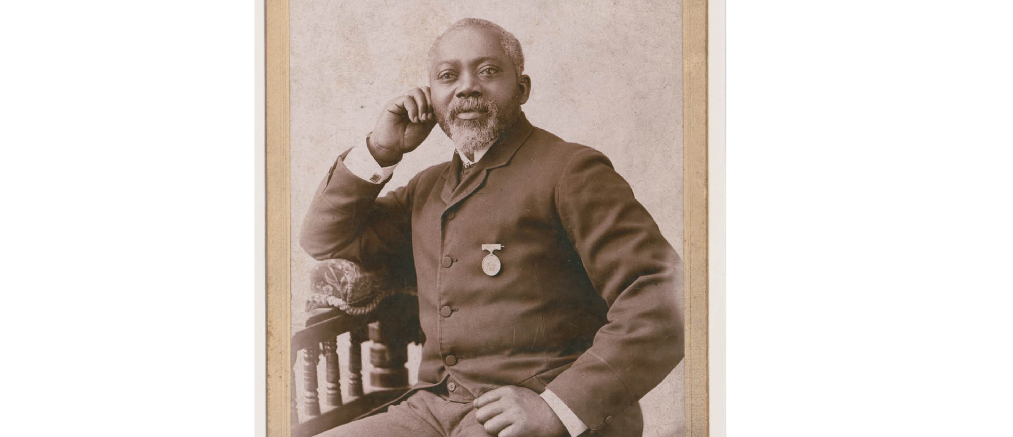 An older Black man, balding with a white beard. He wears a medal on his dress coat. He poses seated, with one hand aside his cheek and the other on his thigh. He looks past the camera. 