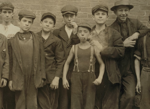 Several children in caps and work clothes pose for the photographer. A small boy, center smokes a pipe. One of his older friends has a good laugh. 