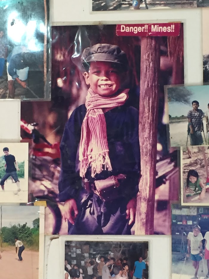 A group of old photos pinned to a wall, in the center, by a sign reading "Danger!! Mines!!", stands a child smiling, eyes shaded by a cap, neck draped in a scarf.