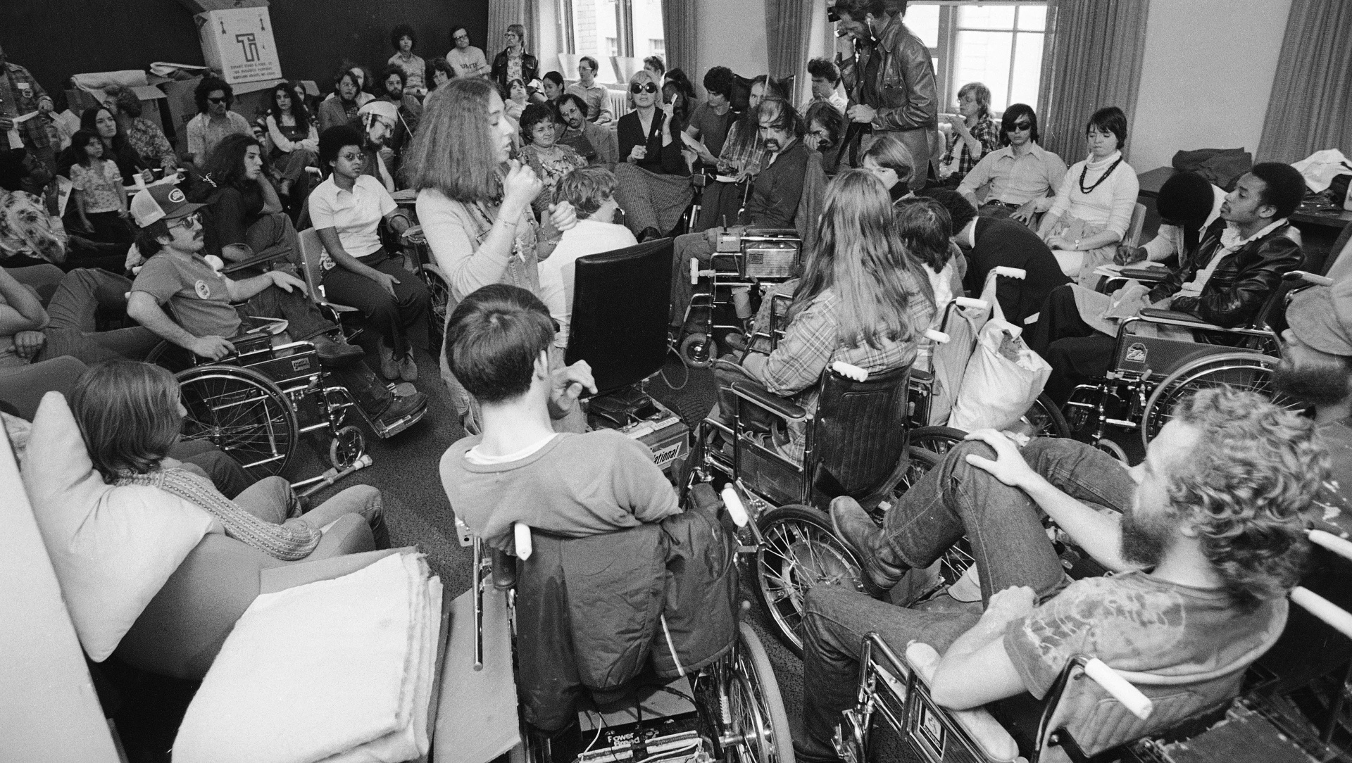 A room filled with organized young people in wheelchairs and other chairs all facing the center, a press camera and tall windows in the background. 