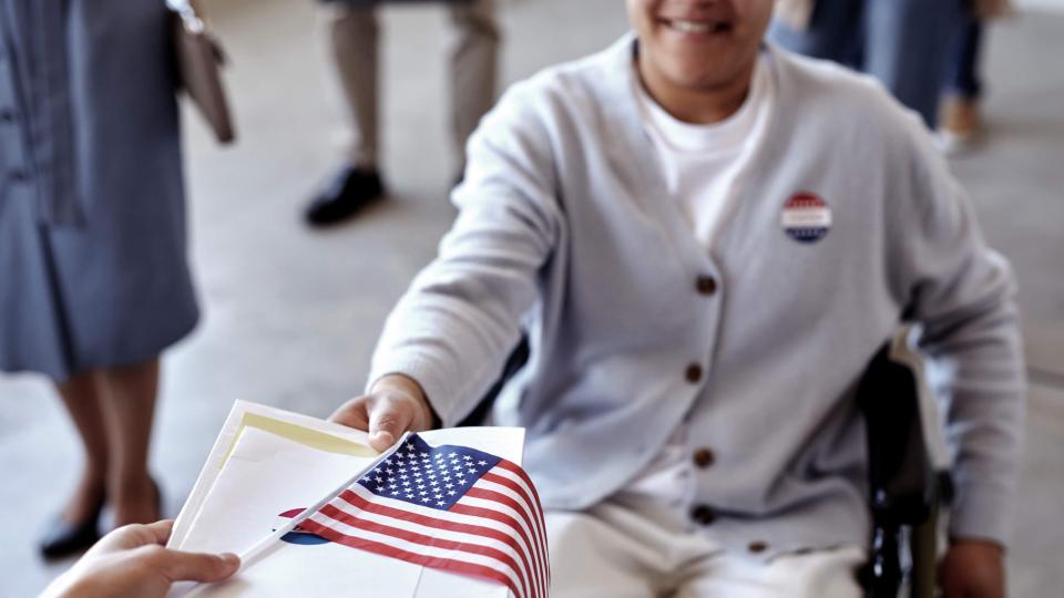 A young smiling person using a wheelchair accepts their ballot, an American flag and an "I voted today" sticker