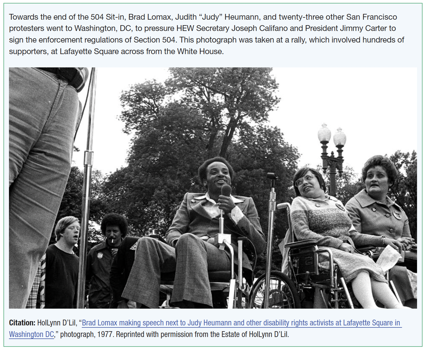 A Black man in a suit holds a microphone and sits in his wheelchair. Beside him a woman with short hair and glasses in a sweater and skirt looks over her shoulder at him from her wheelchair. There are large trees and a fancy lampost behind. Another woman in a suit, also facing the audience, also watches from her wheelchair. A white young man and a Black manwith an Afro stand to one side.