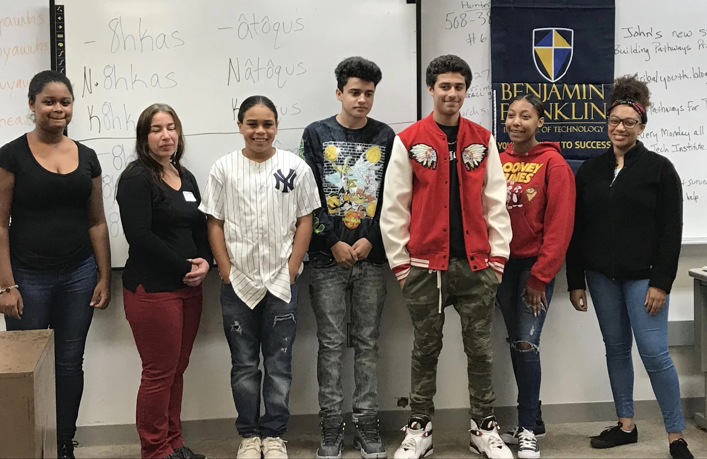 A group of Wampanoag high school students stand with their Indigenous language instructor in front of a white board with written Wôpanâak.
