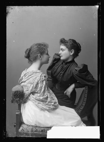 A photograph of Anne Sullivan and Helen Keller. The two women sit on an ornate chair facing each other. Helen, who has her hair in a tight bun and a shawl wrapped around her, leans on the chair arm and is in profile. Anne wears a high collared dress with ling sleeves with her hair in a tight bun, looking at Helen's face. Anne is half standing and half sitting, leaning her elbow on the side of the chair. 