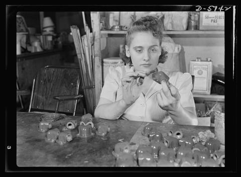 Young woman holds one of dozens of small ceramic pipe fittings at eye level, utility shelves visible behind her. 