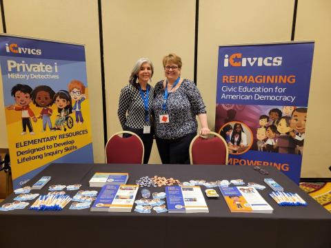 Two teachers stand by posters behind a table at a conference
