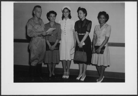 Four smiling African-American women stand smiling as uniformed male soldier stands with them holding an official-looking piece of paper