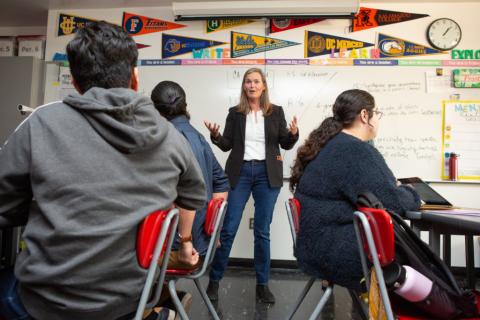 A white woman with long hair a jacket and jeans teaches a class of dark-haired high school students - sports pennants decorate the wall