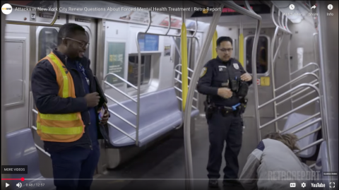 A dark-skinned police officer and a Black man in an orange vest with glasses wake a man with uncombed blonde hair on a New York City Subway train.