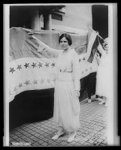 A white woman with dark hair in a bun and wearing a long dress with a sash around the waist holds aloft a glass of wine. Behind her is a banner with two rows of five-pointed stars.