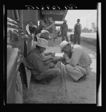 men in overalls and hats squat in the shade of a car as others stand in the background.