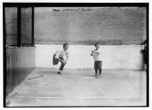 One boy stands on one leg behind a better, on pavement, in front of a brick wall. 