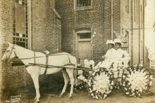 Two young Black women in white hats and long dresses sit in an open carriage with flower-decorated wheels drawn by a white horse in front of a brick building