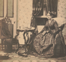 Dorothea Dix in a long dress holds a book and sits in a room with a medical bag on the floor