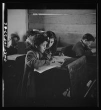 Two girls sharing a single desk in a rustic school lean cheek to cheek, pencils in hand, looking at their schoolwork.
