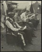 A Native American man in traditional clothes sits with other veterans in a rough encampment.