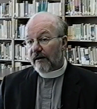 A balding man with a beard and glasses and a priest's collar sits in front of a wall of bookshelves. 