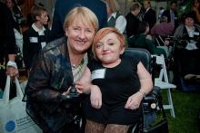 Two women pose outdoors in the midst of a gathering of people of varied abilities and ages. Both women are smiling, and both are white. Stella Young has short red hair swept over one temple. She is of short stature and uses an electric wheelchair. She wears a black dress. Jenny Macklin has short blond hair. She wears a sheer jacket and jacket and kneels by Young.