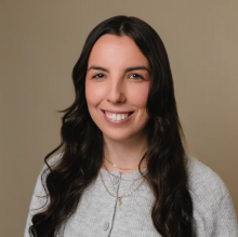 A smiling white woman with long dark hair wears a gold necklace over a sweater.