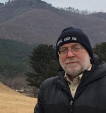 A white man with gray beard and mustache wears a stocking cap and nylon jacket. He stands before a field and trees at the base of a mountain in Korea. 