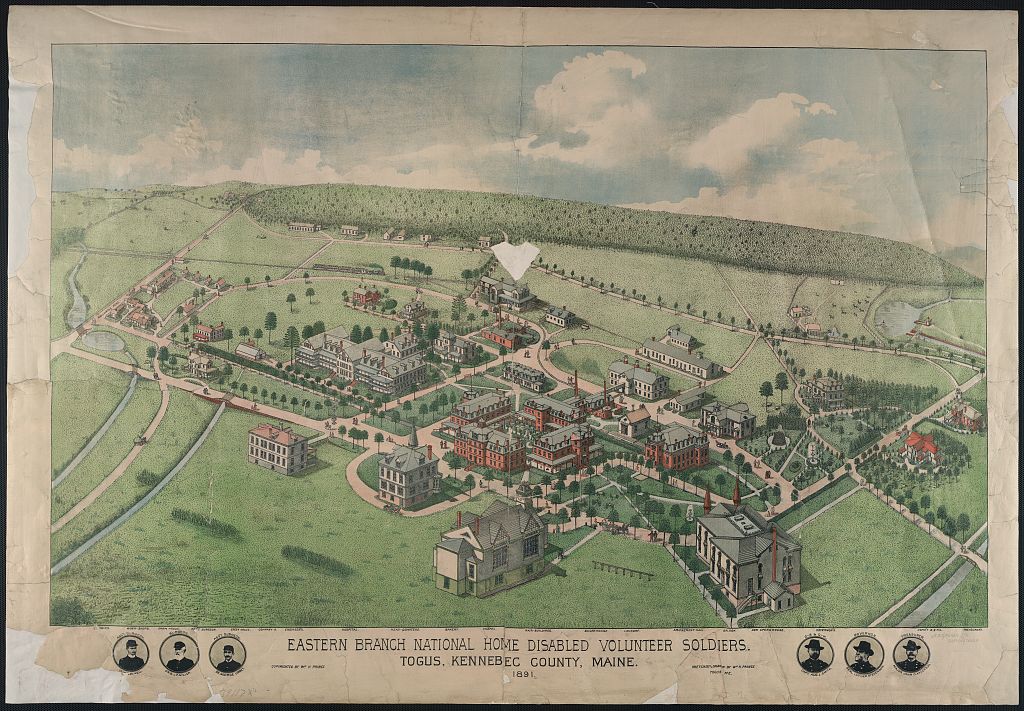 An aerial view shows dozens of large and small wood and brick buildings on a campus with many trees and surrounded by grassy fields.