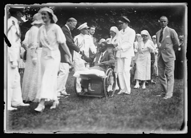 Black and white photo of a lawn where White House dignitaries, including military in "dress whites," and others (possibly President Woodrow Wilson), greeting men in wheelchairs and Red Cross nurses. A black man with a leg elevated in a wooden wheelchair, flanked by a senior officer with decorative braid on his uniform and another man in a dark cap and plain white uniform shakes hands with a white civilian official (President Wilson?). In the foreground, a woman in white with a sash reading "MICHIGAN" walks past a military officer, while behind more nurses and wheelchairs can be seen.