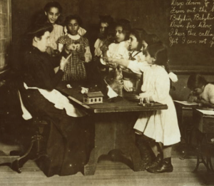 Teacher in high-necked dress and apron, seated at a table with toy house and other objects, surrounded by a group of 6 girls of varied heights, as the littlest girl points and several appear to speak as the teacher points with one hand to a motion her other hand is making.