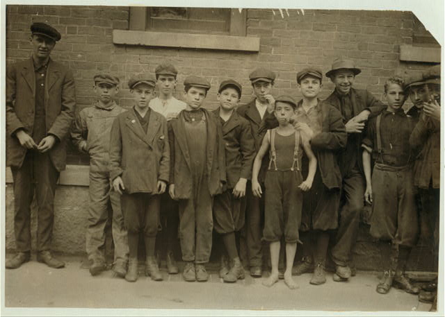 Photo of several boys dressed for factory work. A younger barefoot boy smokes a pipe, probably a prank.
