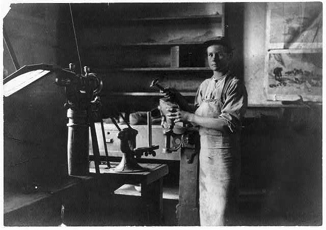 Young man wearing apron stands by machine in factory workshop. 