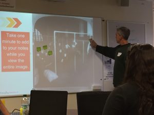 A teacher adds a sticky-note with an imagined thought to a large projected black and white photo on the wall, showing people waiting to have eyes examined at Ellis Island.