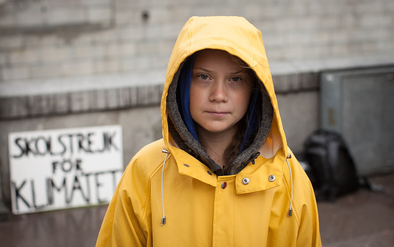 Teenage activist Greta Thunberg stands outside wearing a hooded yellow raincoat, "Skolstrekt for Klimatet" sign behind her [school strike for climate]