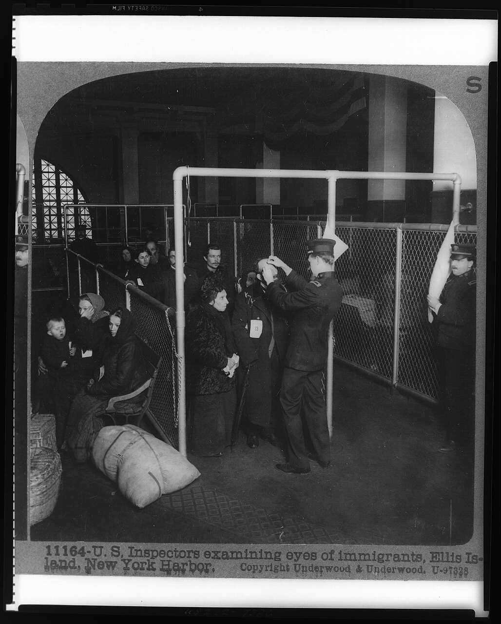 Old photo of people with bundles lined up for inspection in a high-ceilinged hall lit by an enormous window in the distance. "