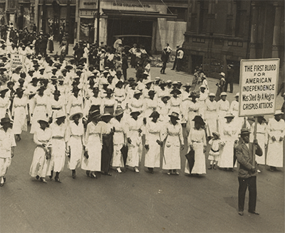 thousands of black women wearing white long dresses and some men in suits march in a 1917 New York City street. Three protest signs are visible. 
